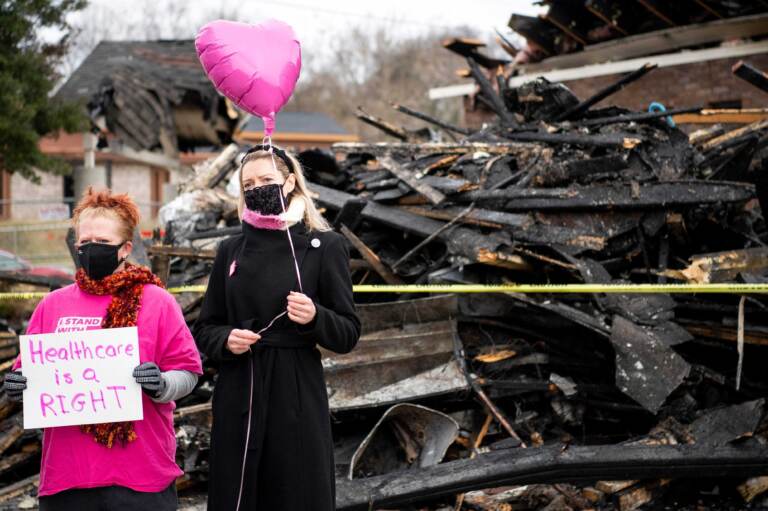 Two people holds a pink balloon in front of a pile of burnt wreckage.