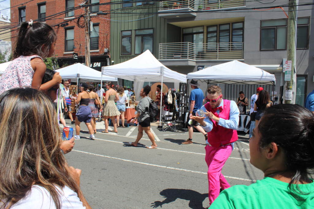 A street performer dressed in a pink suit displays a magic ball as spectators look on. Vendors are set up underneath white tents in the background as people walk by.