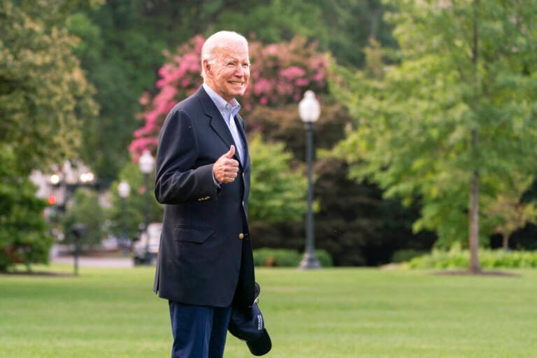 President Biden smiles and gives a thumbs up sign. Trees and grass are visible in the background.