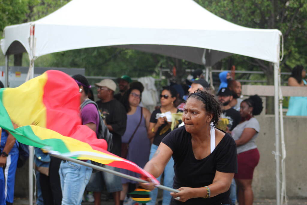 A woman waves the Rastafarian flag, with a white tent and crowd of people visible in the background.