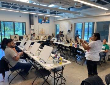 A person speaks and gestures at the front of a room, where students are sitting before blank easels.