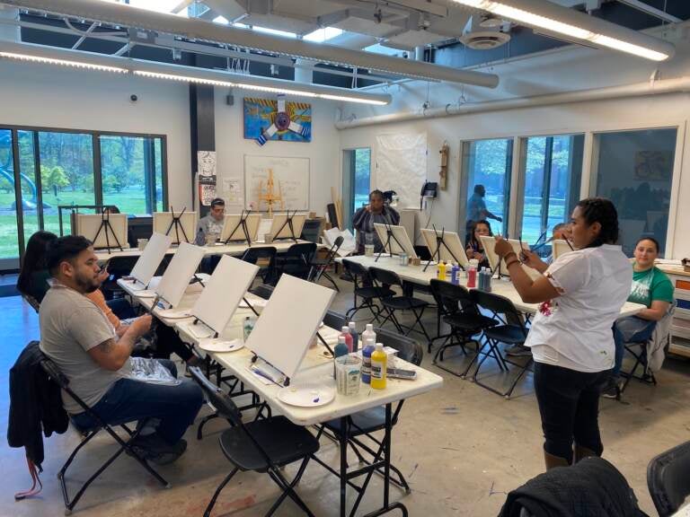 A person speaks and gestures at the front of a room, where students are sitting before blank easels.