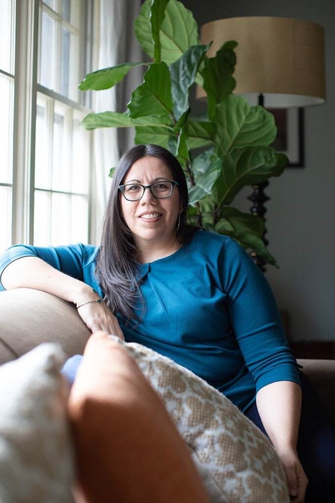 A woman sits with her arm on the back of a sofa, next to a window. A plant hangs in the background.