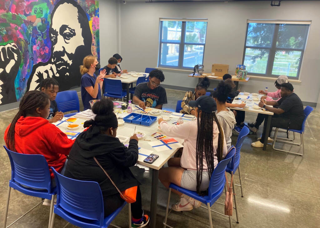 A group of students are seated in blue chairs around rectangular tables with art supplies in front of them. The view is from above.