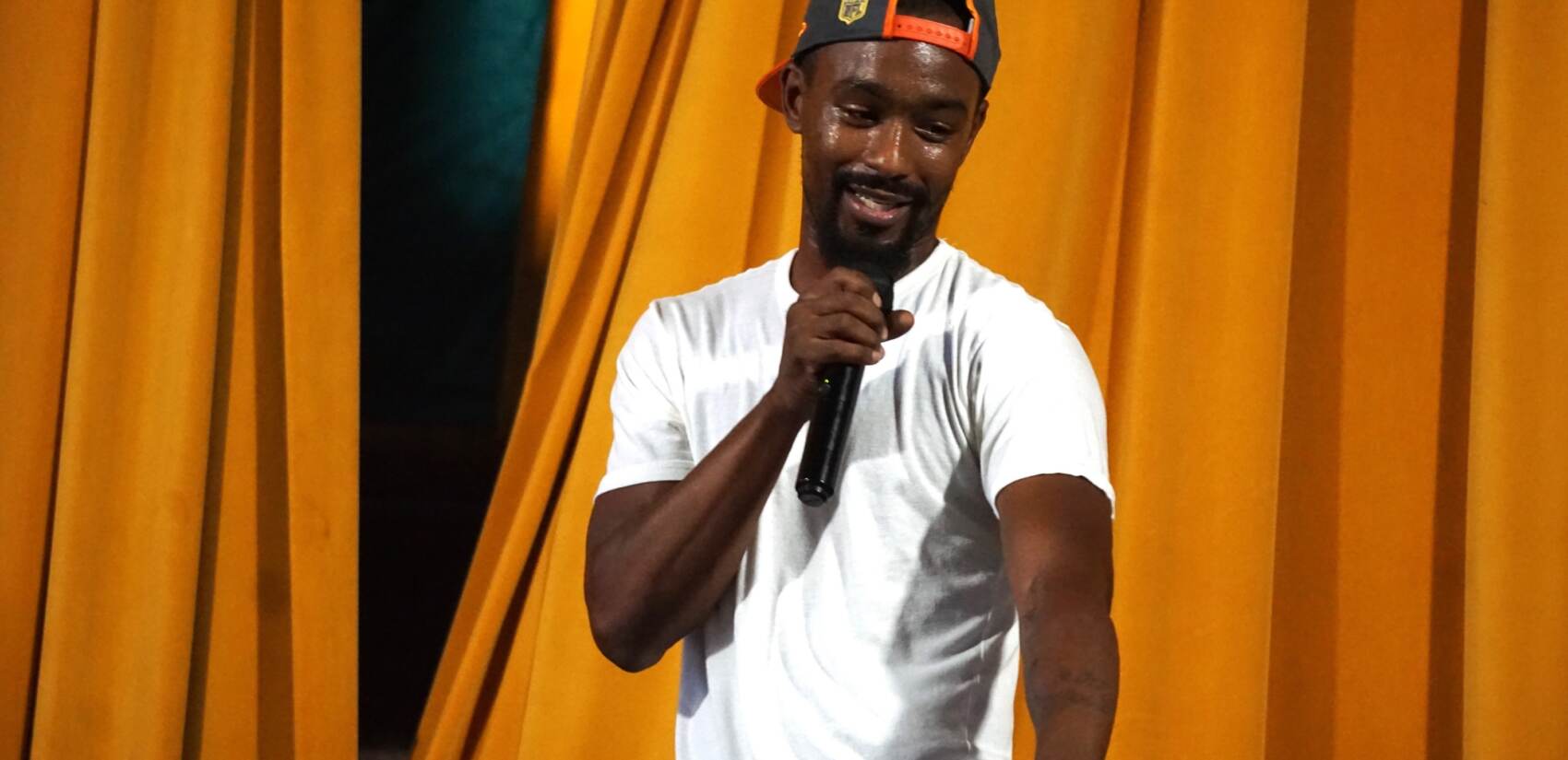 A person holds a microphone and gestures. Stage curtains are visible in the background.