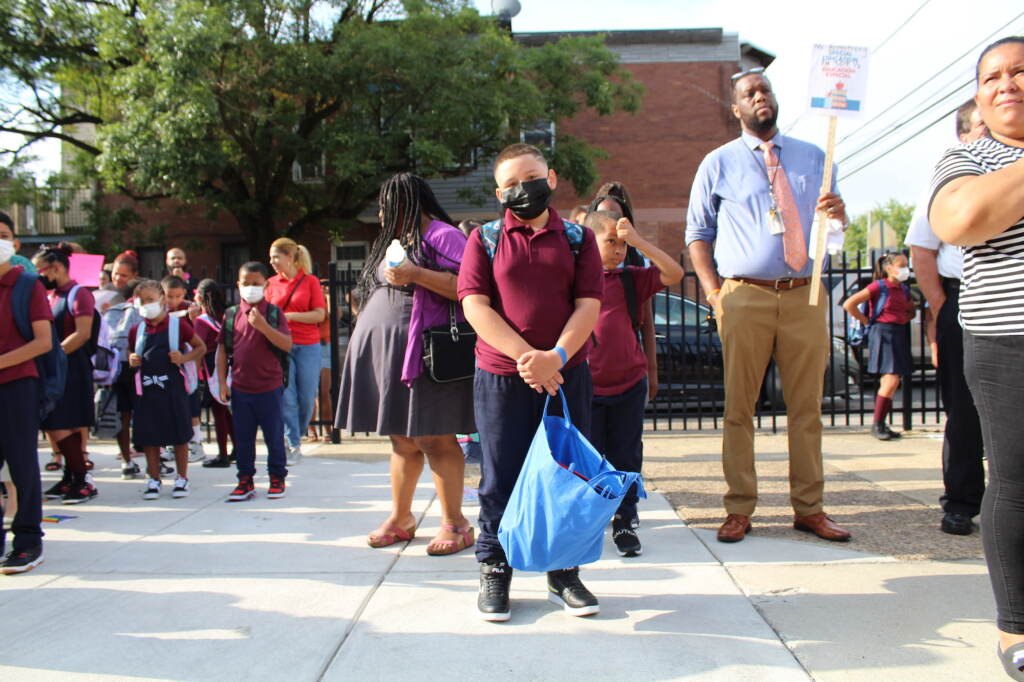 A student in a red polo shirt holds a blue bag, as other students and teachers stand behind him.