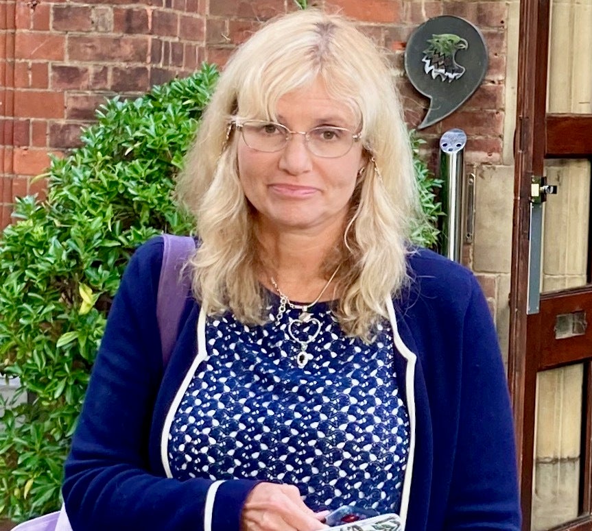 Headshot of a woman with glasses, with a brick building and bushes in the background.