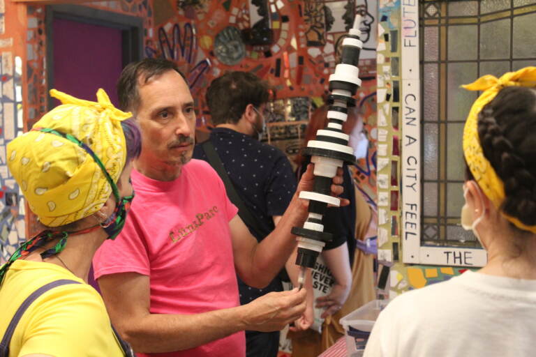 The artist, in a pink T-shirt in the center, holds up plastic recycled art while two people, backs to the camera, listen.
