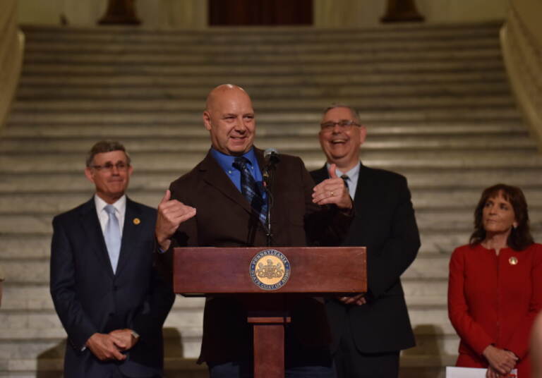 Mastriano speaks and gestures at a podium. Other men stand in the background.