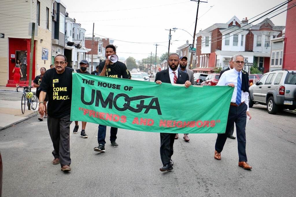 A group of men walk down a street bordered by rowhomes with a sign that reads "The House of Umoja Friends and Neighbors."