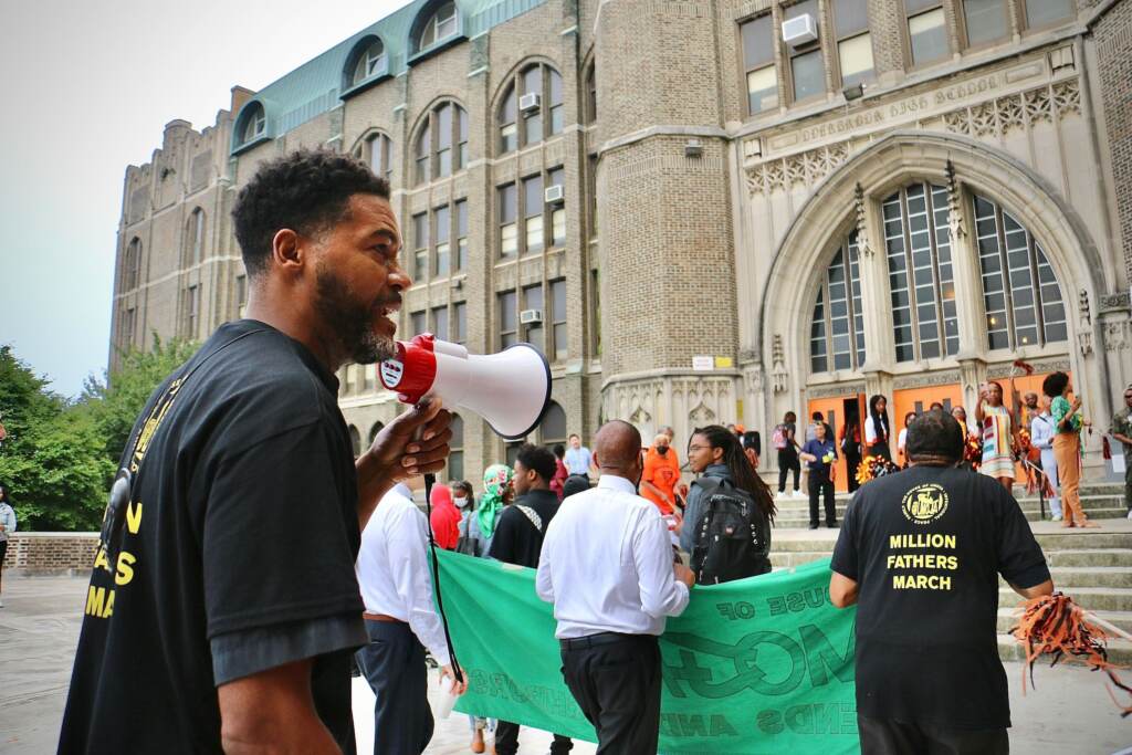 A man with a megaphone stands in front of the entrance to a high school, with other people standing behind him.