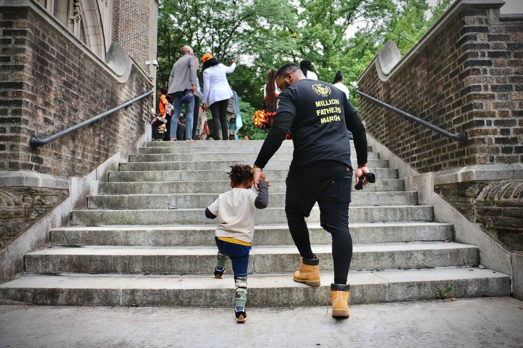 A father holds his son's hand as they both walk up a set of stairs.