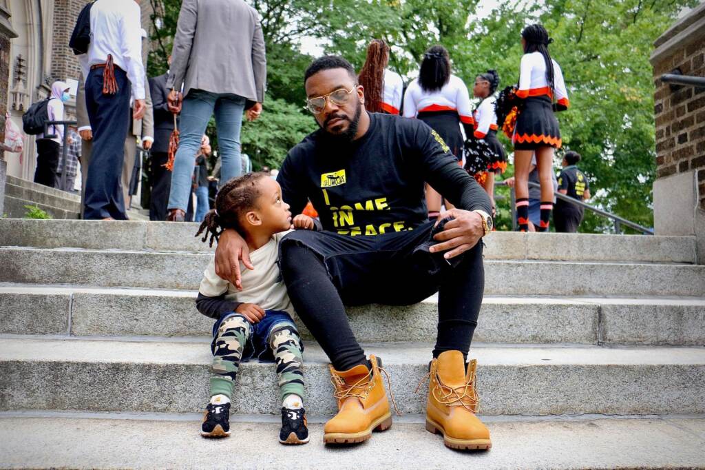 A father sits on steps with his arm around his young son, who is turned towards him and looking up at him.