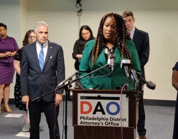 A woman in a green blouse speaks at a podium with a sign in front which reads DAO. People stand in the background.