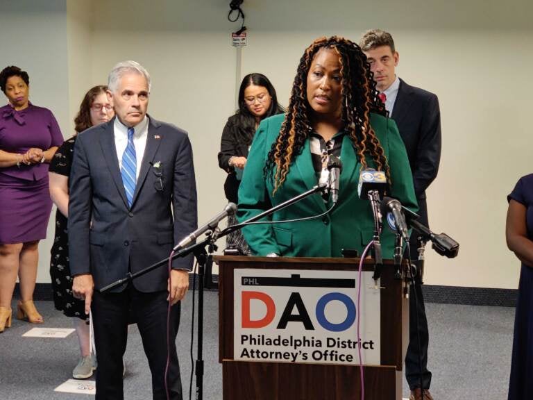 A woman in a green blouse speaks at a podium with a sign in front which reads DAO. People stand in the background.