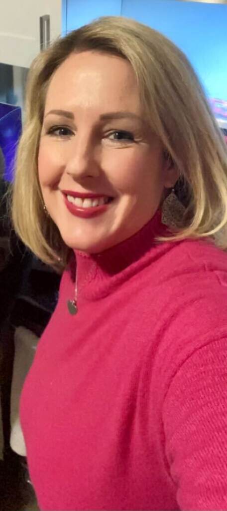 A woman smiles into the camera, with blue sky visible behind her.