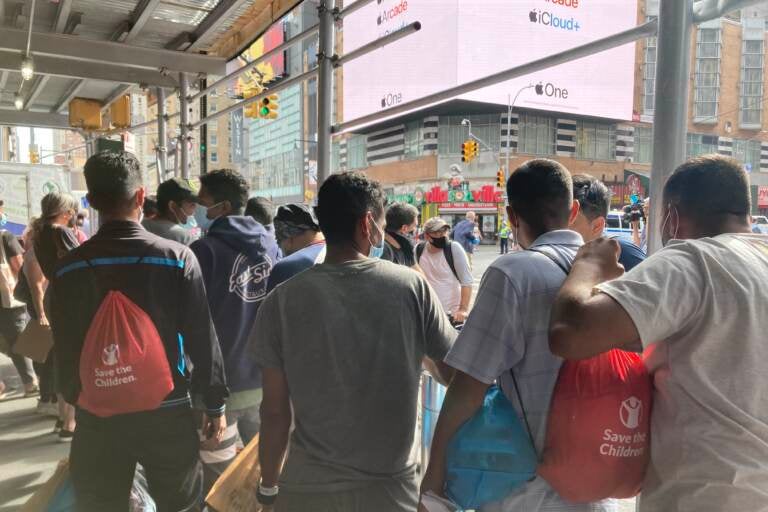 A group of migrants arriving in New York City are shown from behind as they look out on a city street.