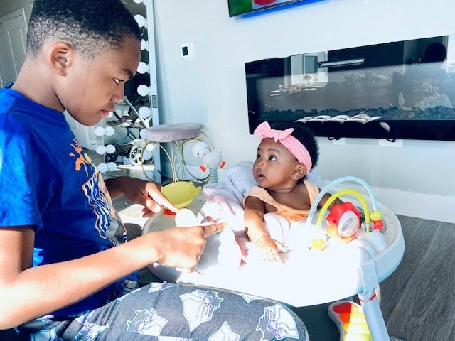 A boy plays with his little sister, who is sitting in a high chair.