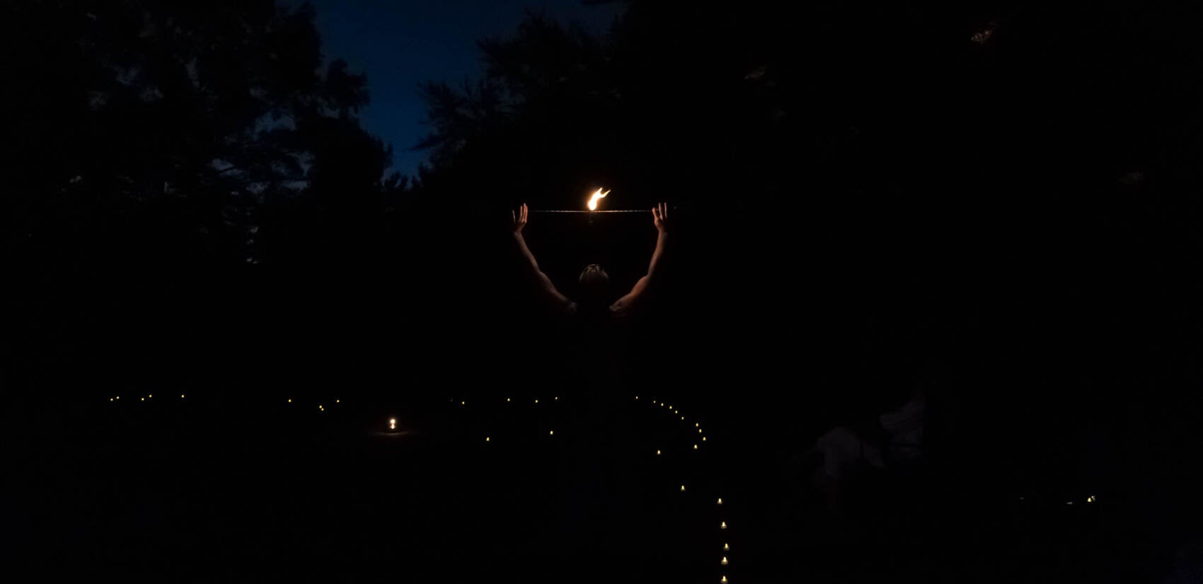 A man holds up a lighted fire spinner, as all around him is dark.