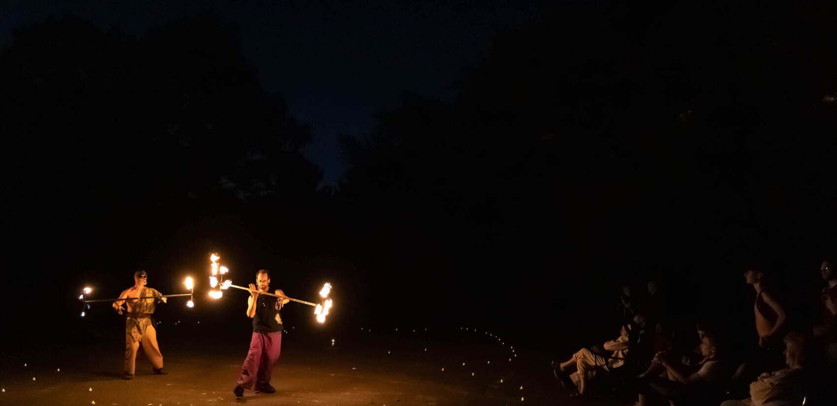 Two people perform fire spinning inside of a circle as a crowd of people looks on at the right.