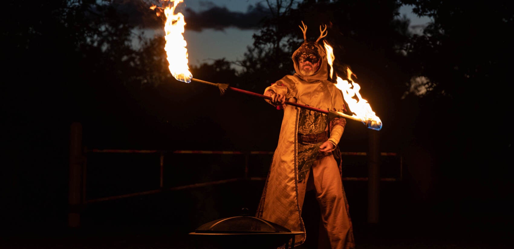 A man in an antler suit performs fire-spinning, with the last rays of sunlight fading behind him.