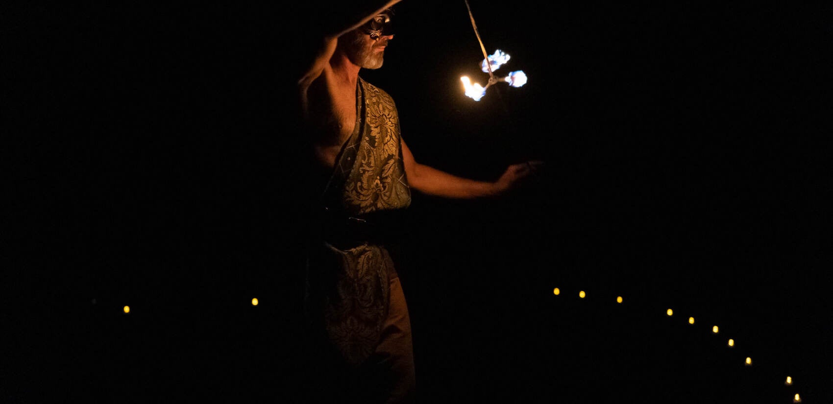 A man holds a lit fire-spinner in front of him in the dark.