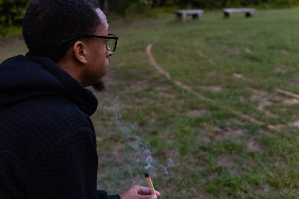 A close-up of a young man carrying palo santo, with green grass and benches visible in the background.