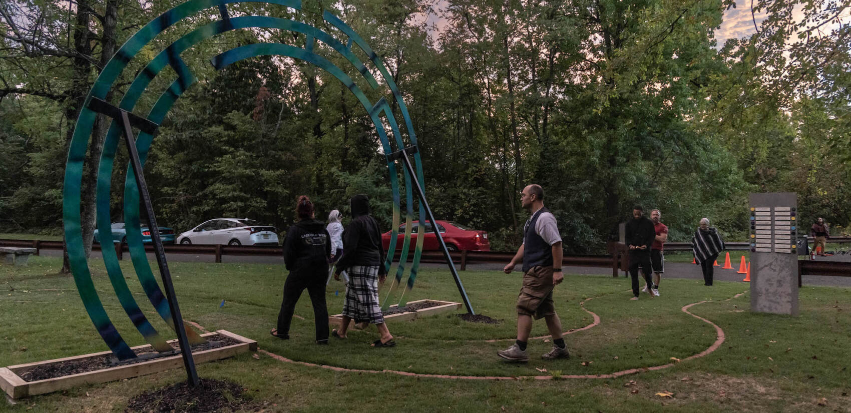 People walk in a green park at sunset, some walking under a sculpted arch.