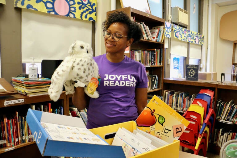 A woman in a purple shirt holds a stuffed owl in a classroom.