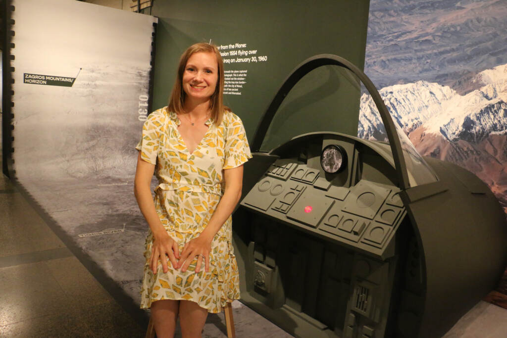 A woman sits, smiling, posing in front of an exhibition.