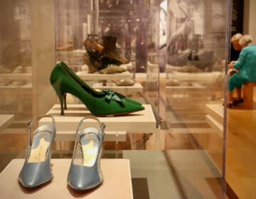 A pair of green high heels and silver high heels are in a glass display case. Museum-goers are visible in the background.