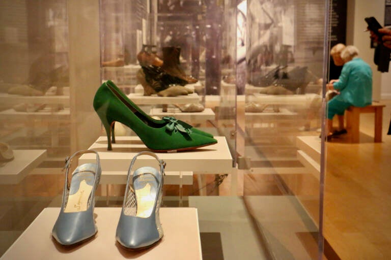 A pair of green high heels and silver high heels are in a glass display case. Museum-goers are visible in the background.