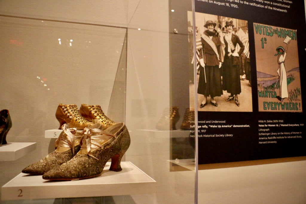 Shoes are shown in a glass display case, with a sign with information about the suffragette movement visible in the background.