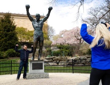 In this April 3, 2020, photo, Jessica Shiroff, right, directs exchange student Joao Martucci, of Brazil, as he poses for a photograph with the Rocky Statue at the Philadelphia Art Museum in Philadelphia. “Rocky” finished tied for No. 2 in The Associated Press Top 25 favorite sports movies poll. (AP Photo/Matt Rourke)