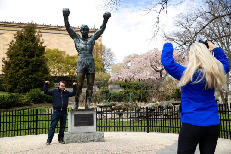 In this April 3, 2020, photo, Jessica Shiroff, right, directs exchange student Joao Martucci, of Brazil, as he poses for a photograph with the Rocky Statue at the Philadelphia Art Museum in Philadelphia. “Rocky” finished tied for No. 2 in The Associated Press Top 25 favorite sports movies poll. (AP Photo/Matt Rourke)