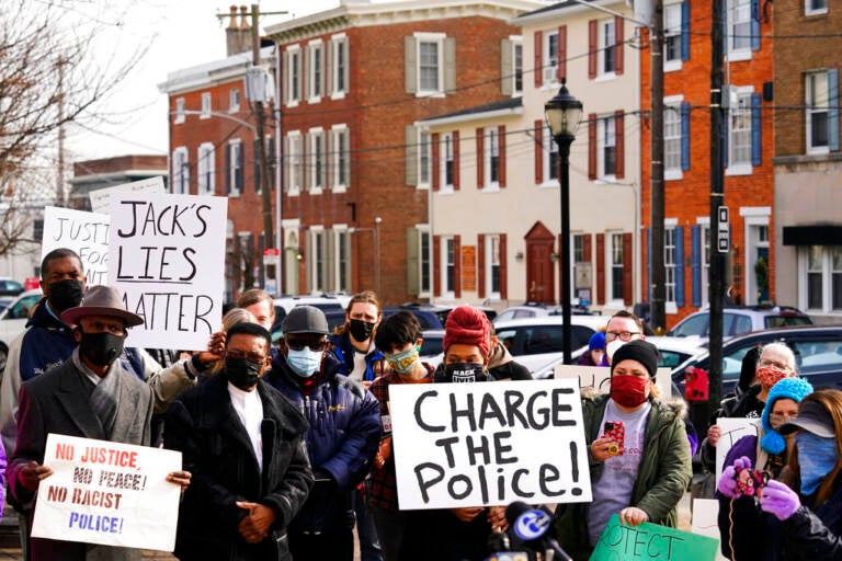 People gather holding signs that read Charge the police. Rowhomes are visible in the background.