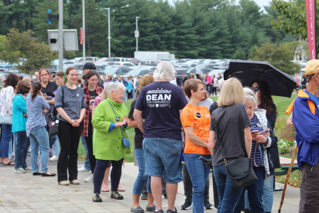 A line of people stands outside. Trees are visible in the background, along with a parking lot.