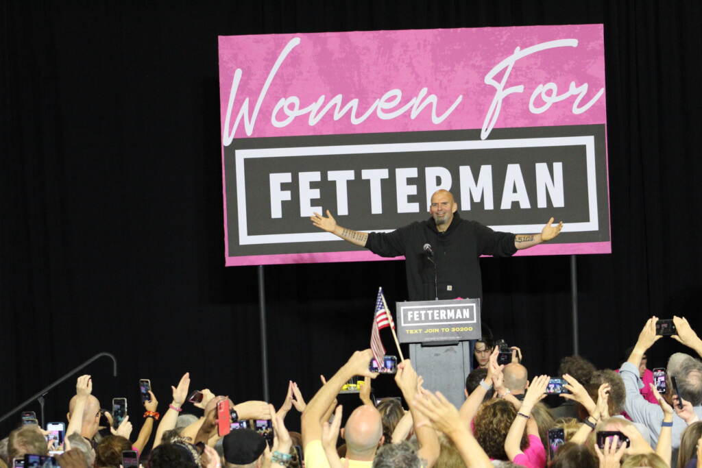 John Fetterman stands at a podium with arms outraised, with people cheering in front of him. The sign behind him reads, "Women for Fetterman."