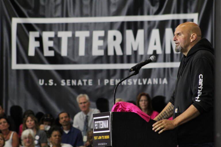 Fetterman is visible in profile, speaking at a podium in front of a crowd. A huge Fetterman campaign sign hangs in the background.