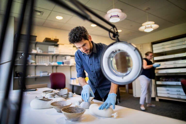 Researcher Simcha Gross lays out incantation bowls in the research room at the Penn Museum, nestling the objects atop long bags filled with silica gel for support. In the background, museum object keeper Katherine Blanchard pulls an item from the storage cabinet. (Eric Sucar/Penn Museum)