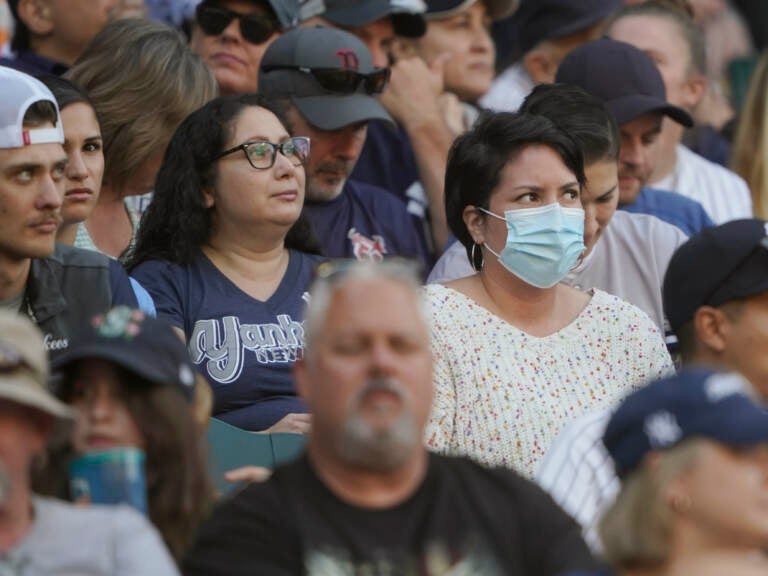 A crowd of people are visible, most not wearing masks, one person wearing a blue surgical mask.