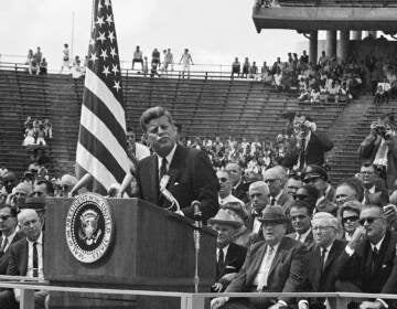 A black-and-white photo of President Kennedy speaking in the midst of a large crowd.