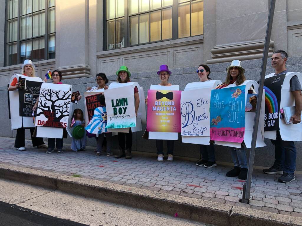 A group of people wearing signs representing banned books stand on a sidewalk.