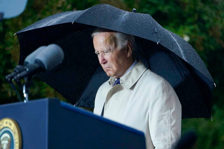 President Biden stands under a black umbrella at a podium, with a somber expression on his face.