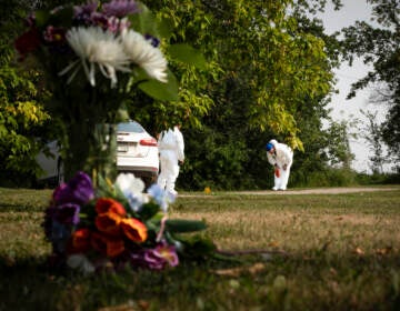 A close-up of flowers on the ground, with an investigator examining a crime scene in the background.