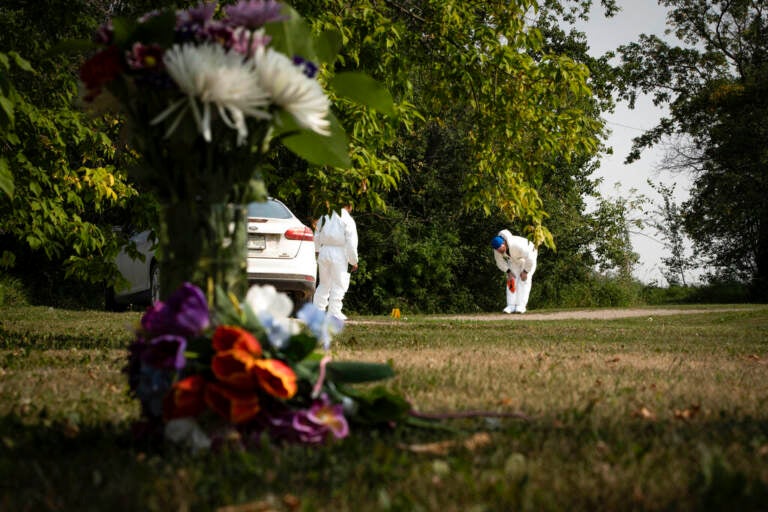 A close-up of flowers on the ground, with an investigator examining a crime scene in the background.