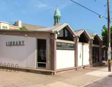 A white building with arches is visible on a street corner on a sunny day. Green trees are on the left and right side of the photo.