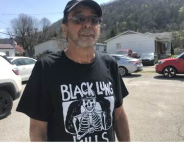 A man with a baseball hat and sunglasses faces the camera. He's wearing a shirt that reads, 