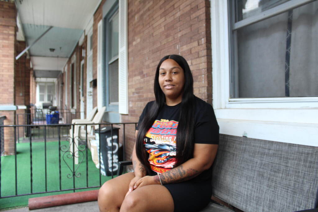 A woman sits on a couch on the porch of a rowhome. She is smiling and her hands are folded in her lap.