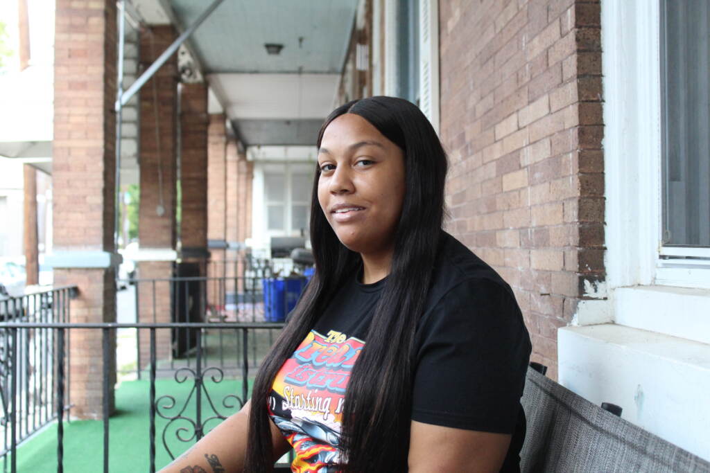 A woman sits on her porch, looking towards the camera and slightly smiling.